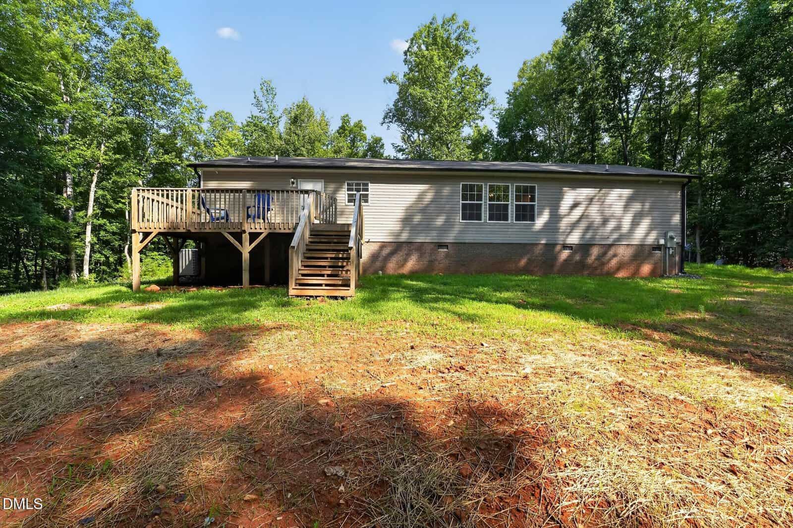 Single-story house with beige siding and brick foundation, a wooden deck with stairs, and surrounded by trees. The yard has patches of bare red soil and grass.