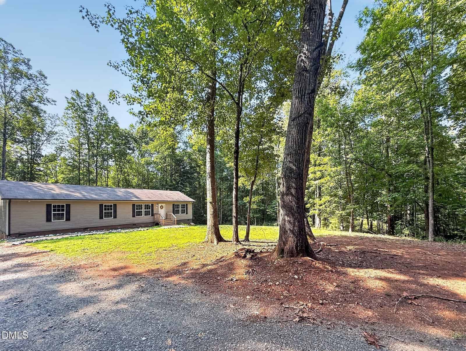 Single-story beige house with a front porch, surrounded by tall trees and a gravel driveway, on a sunny day. The area around the trees has exposed soil and patches of grass.