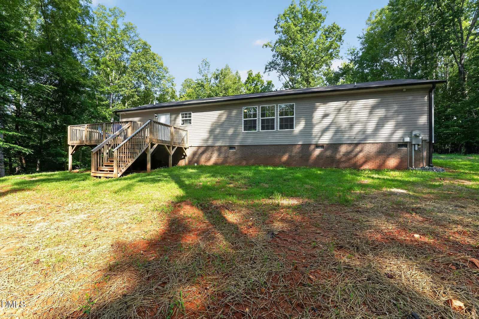 Single-story beige house with a brick foundation, wooden deck, and stairs, surrounded by grass and trees on a sunny day.