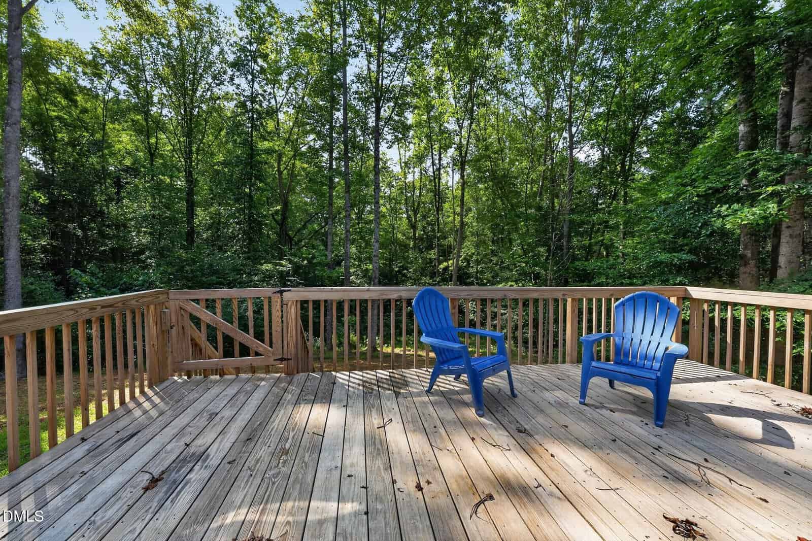A wooden deck with two blue plastic chairs overlooks a lush, green forest. Sunlight filters through the tall trees, casting shadows on the deck.