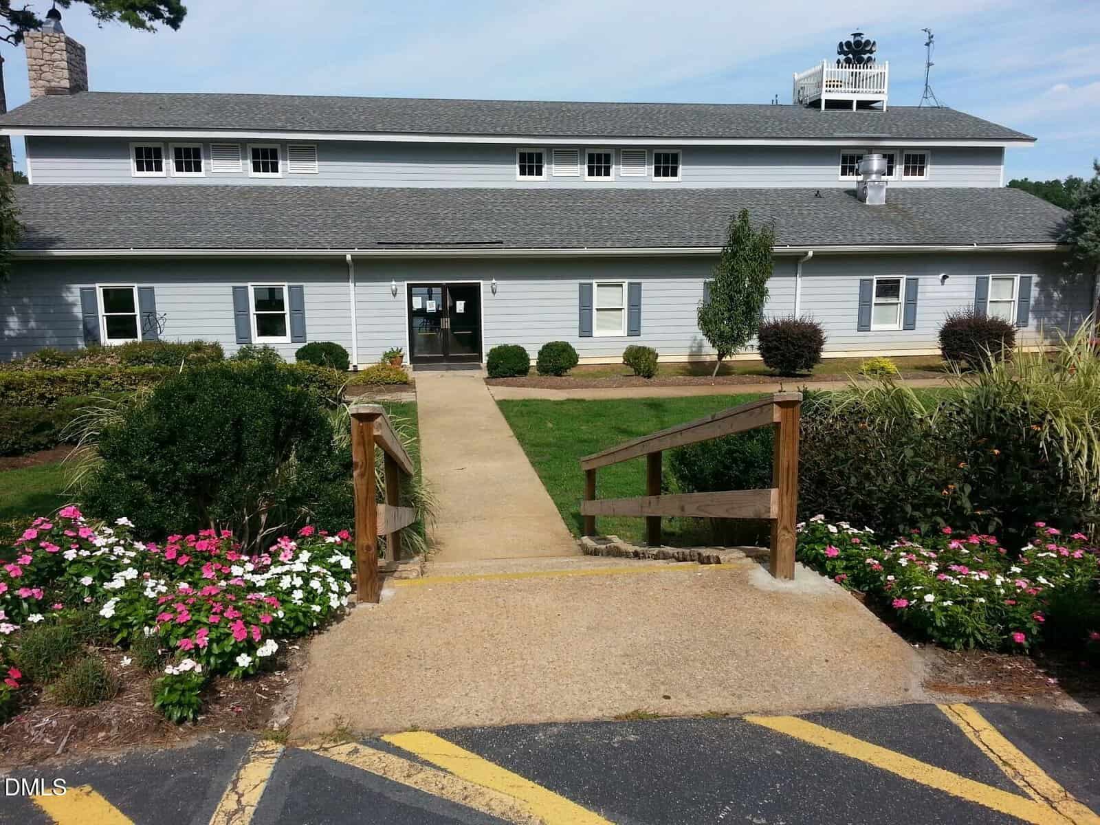 A long, light gray building with many windows and a central entryway. A path leads up to the door, bordered by grass and blooming flowers, with wooden railings along the steps in the foreground.