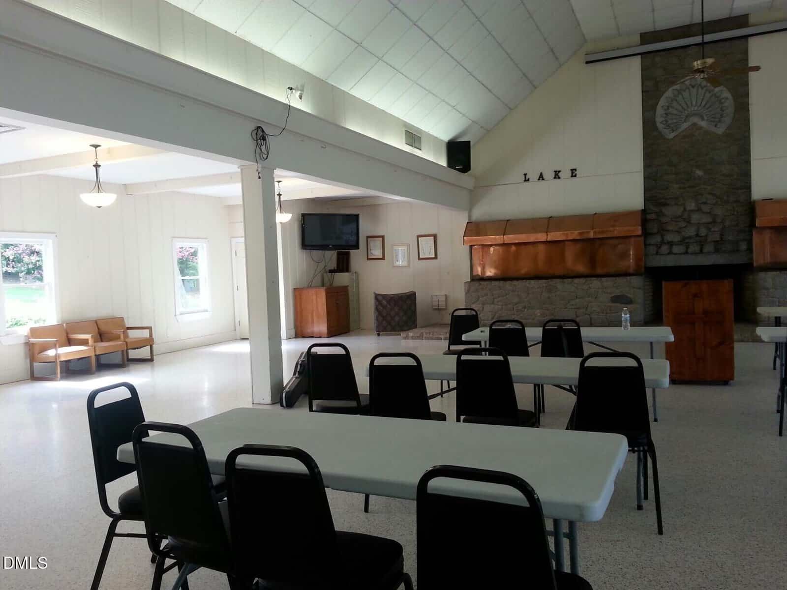 A spacious room with white walls and ceiling, several rectangular tables with black chairs, a stone fireplace, copper hood, and a mounted TV. Natural light enters from windows and doors on the left.