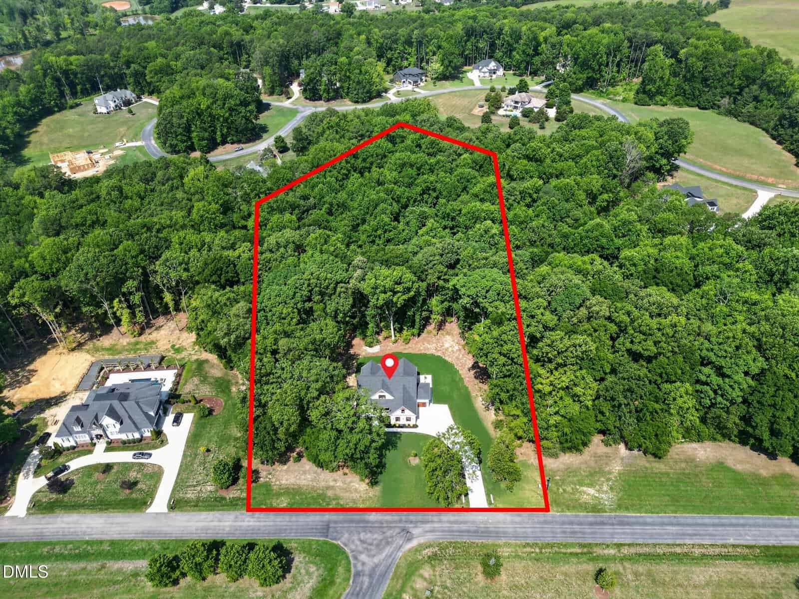 Aerial view of a house surrounded by trees, with a red outline marking its large lot. The home sits near a road, with neighboring houses and forested areas nearby. A location pin points to the house.