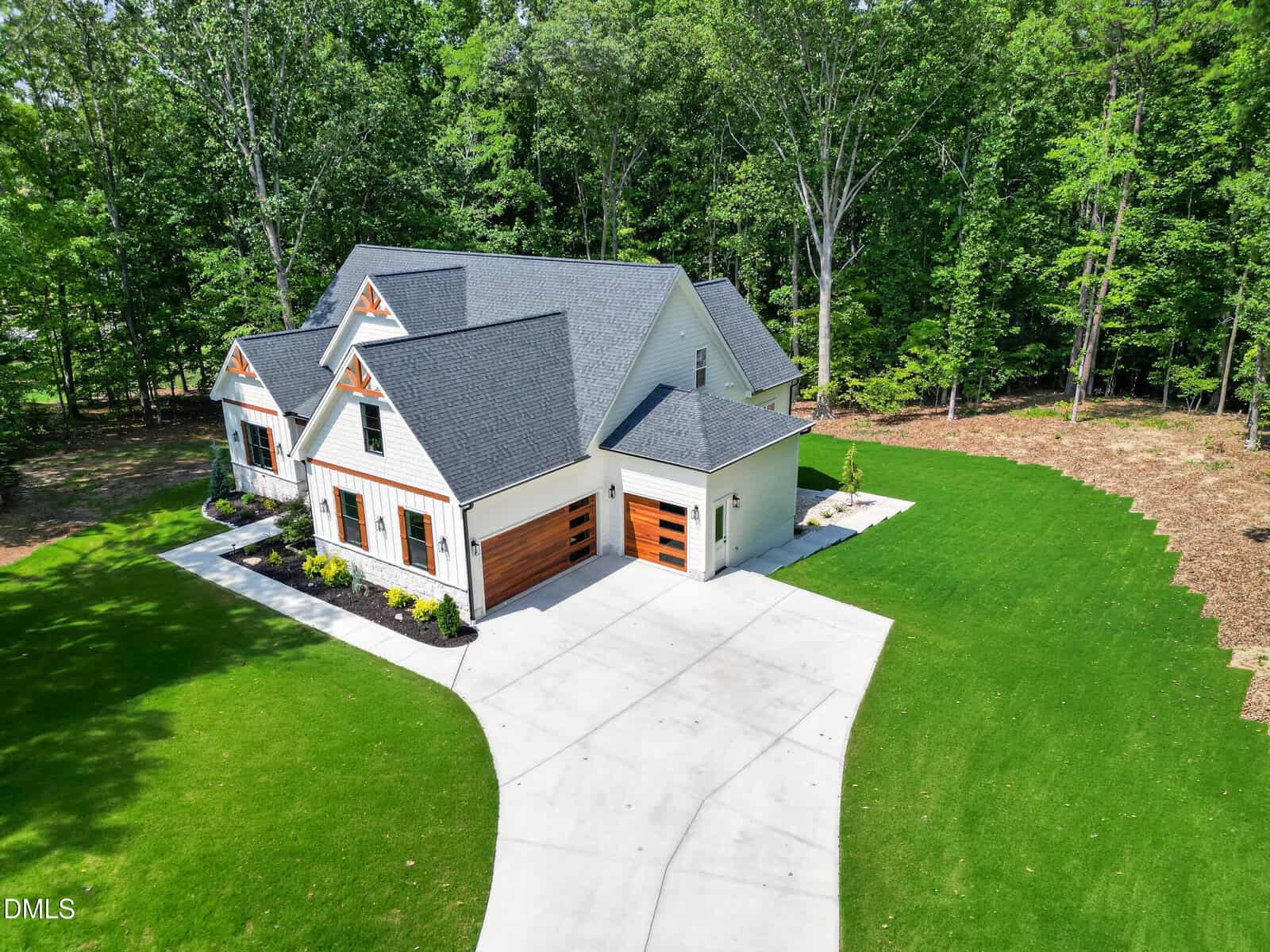 A modern white house with wood accents, three garage doors, and a large driveway, surrounded by green lawn and tall trees under a bright sunny sky.