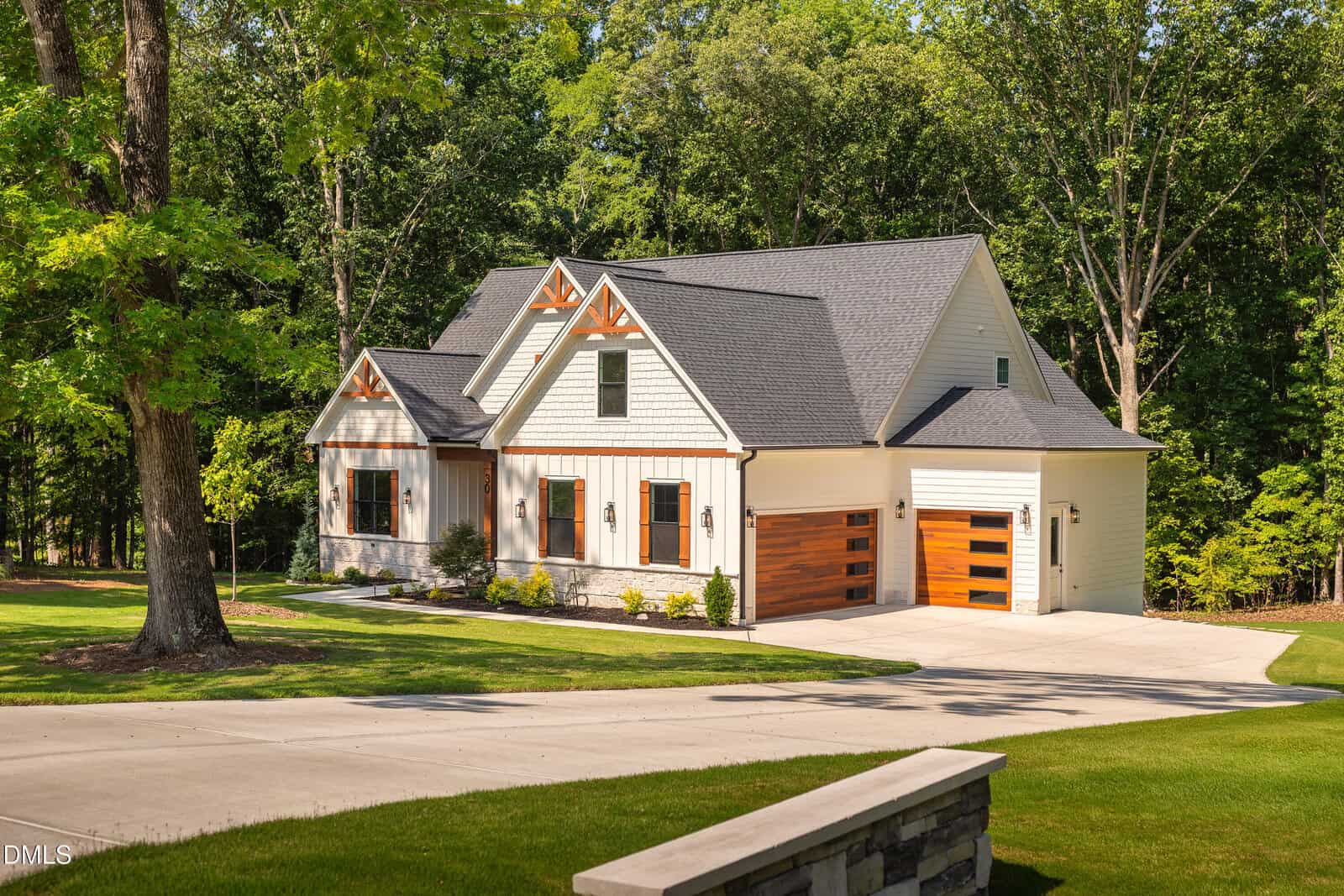 A modern farmhouse at 30 Park Meadow Lane in Youngsville, with white siding, wood accents, and a three-car garage, is surrounded by green grass, tall trees, and a curved driveway on a sunny day.
