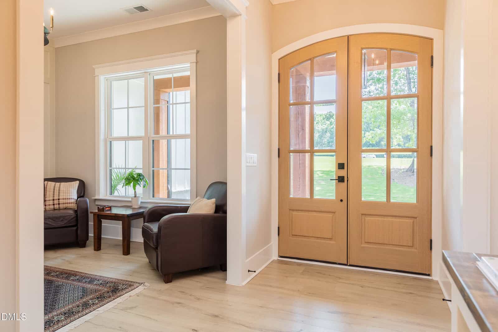 Bright entryway with double wooden front doors, two dark armchairs, small side table, potted plant, large window, and light wood floors. Sunlight streams into the space, adding warmth and inviting atmosphere.