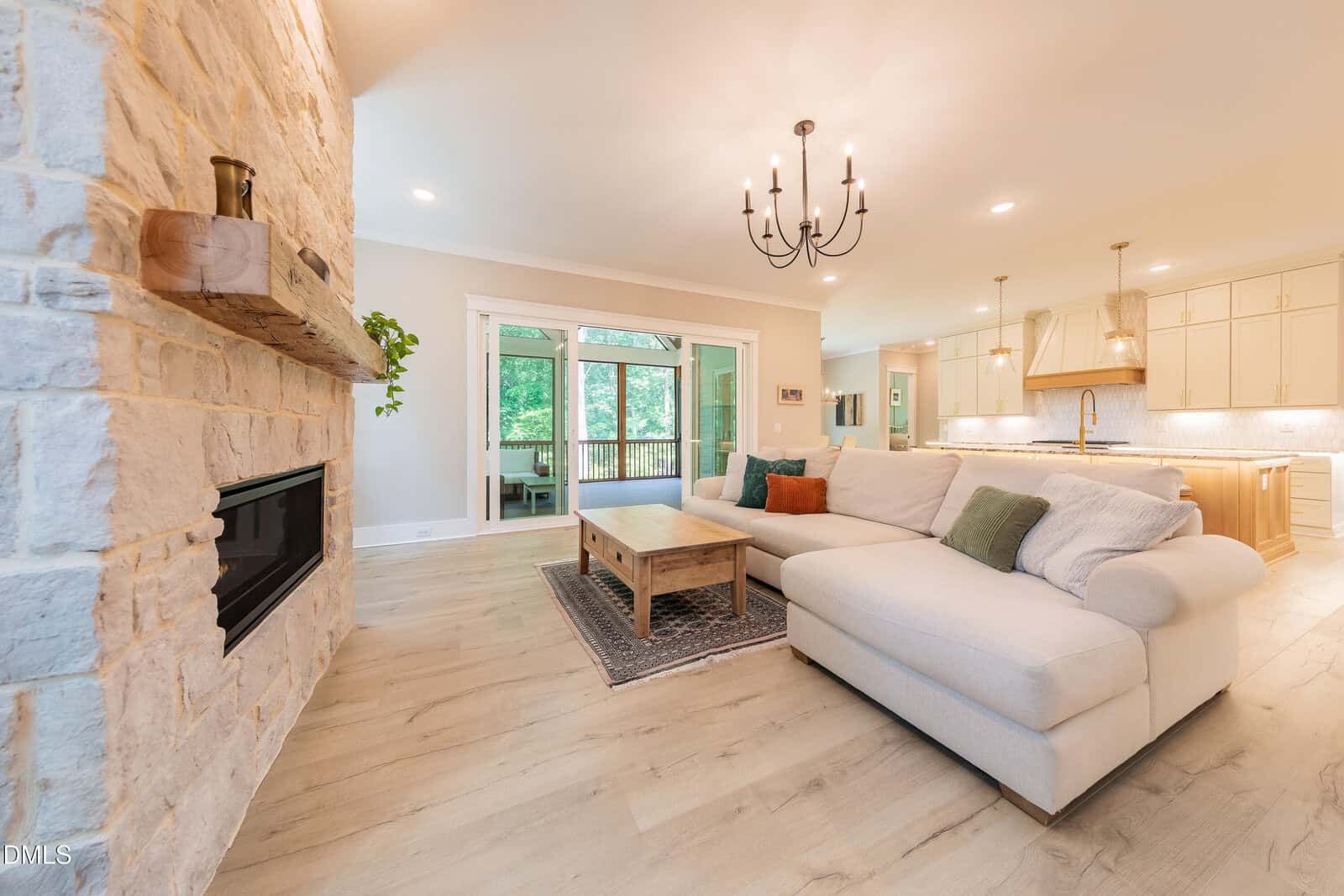Bright living room with light wood floors, a stone fireplace, white sectional sofa with colorful pillows, wooden coffee table, and an open kitchen in the background. Large glass doors reveal a green outdoor view.