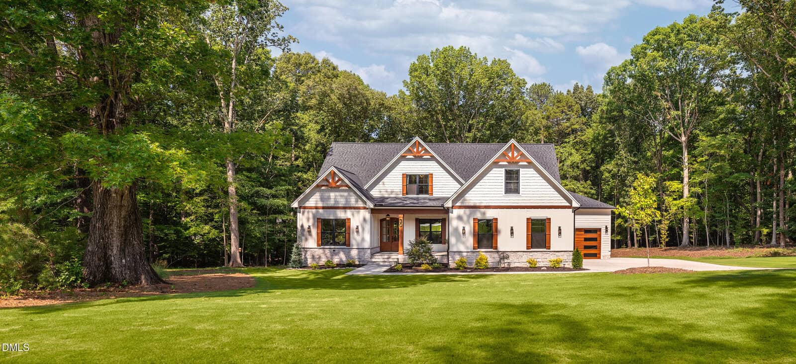 A modern cottage-style house with a gray roof, white siding, and wood accents sits on a large green lawn, surrounded by tall trees under a partly cloudy sky.