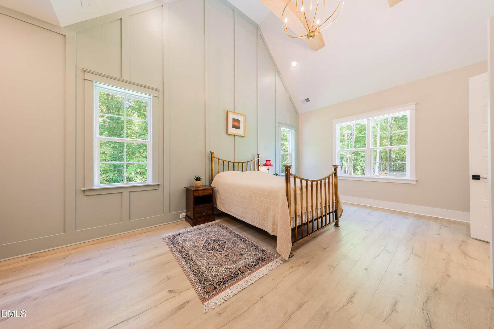 Bright bedroom with wood floors, a single bed, nightstand, small rug, and large windows letting in natural light. Light-colored walls and vaulted ceiling with chandelier complete the spacious look.