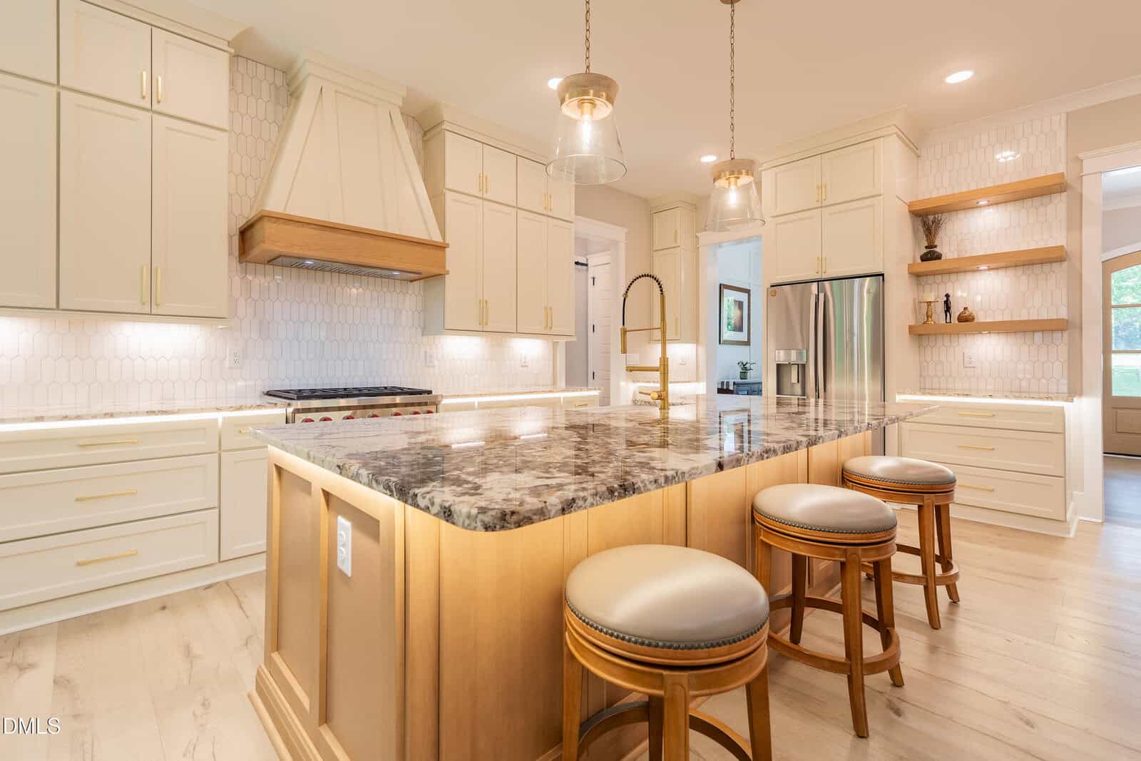 Modern kitchen with cream cabinets, wood accents, marble island with sink, three round stools, stainless steel appliances, open shelves, pendant lights, and light wood flooring.