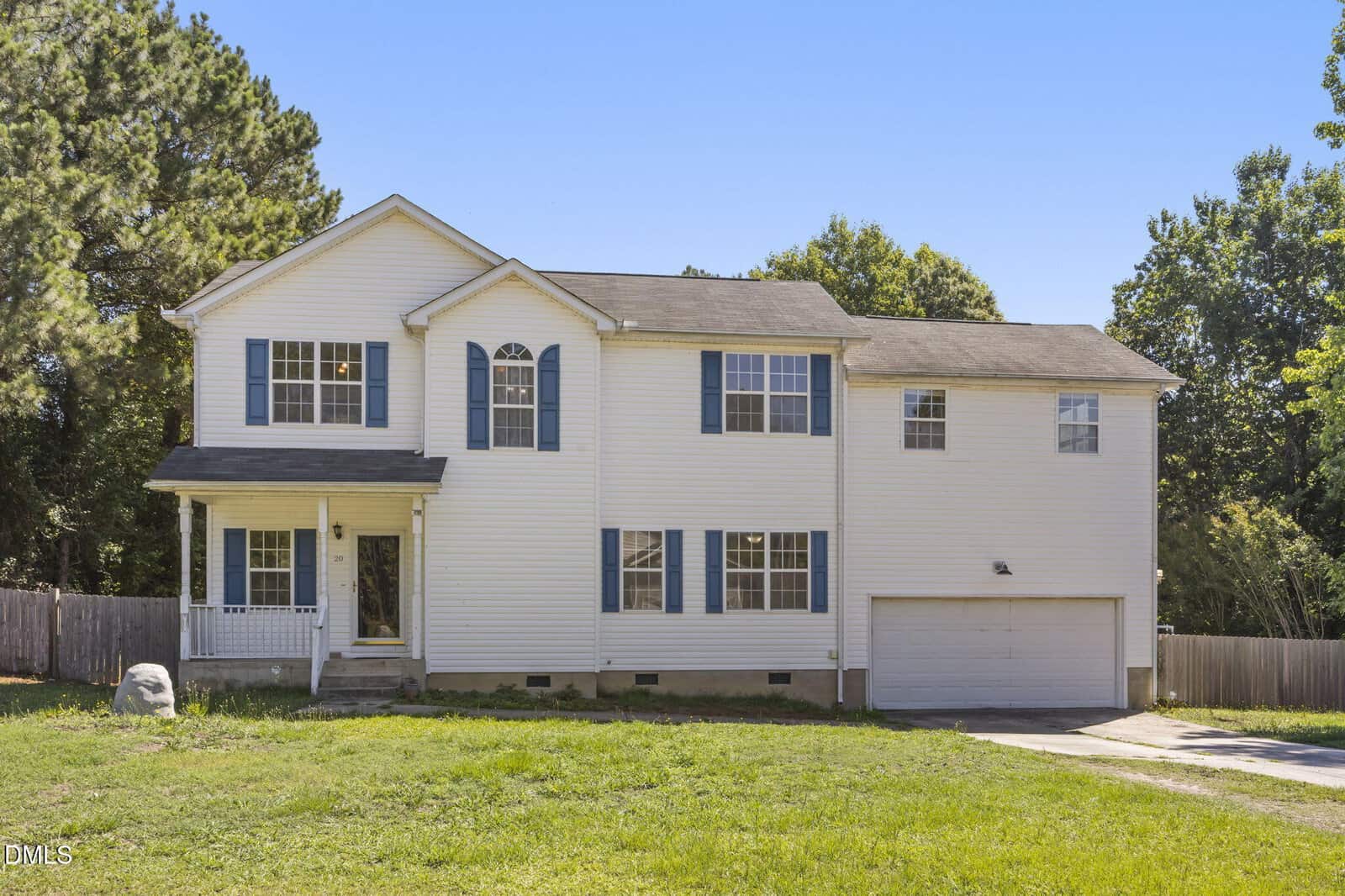 A two-story white house with blue shutters, a covered porch, attached two-car garage, and a large green lawn in front invites you to 20 Greenleaf Court in Louisburg. Trees and a wooden fence are visible in the background under a clear blue sky.
