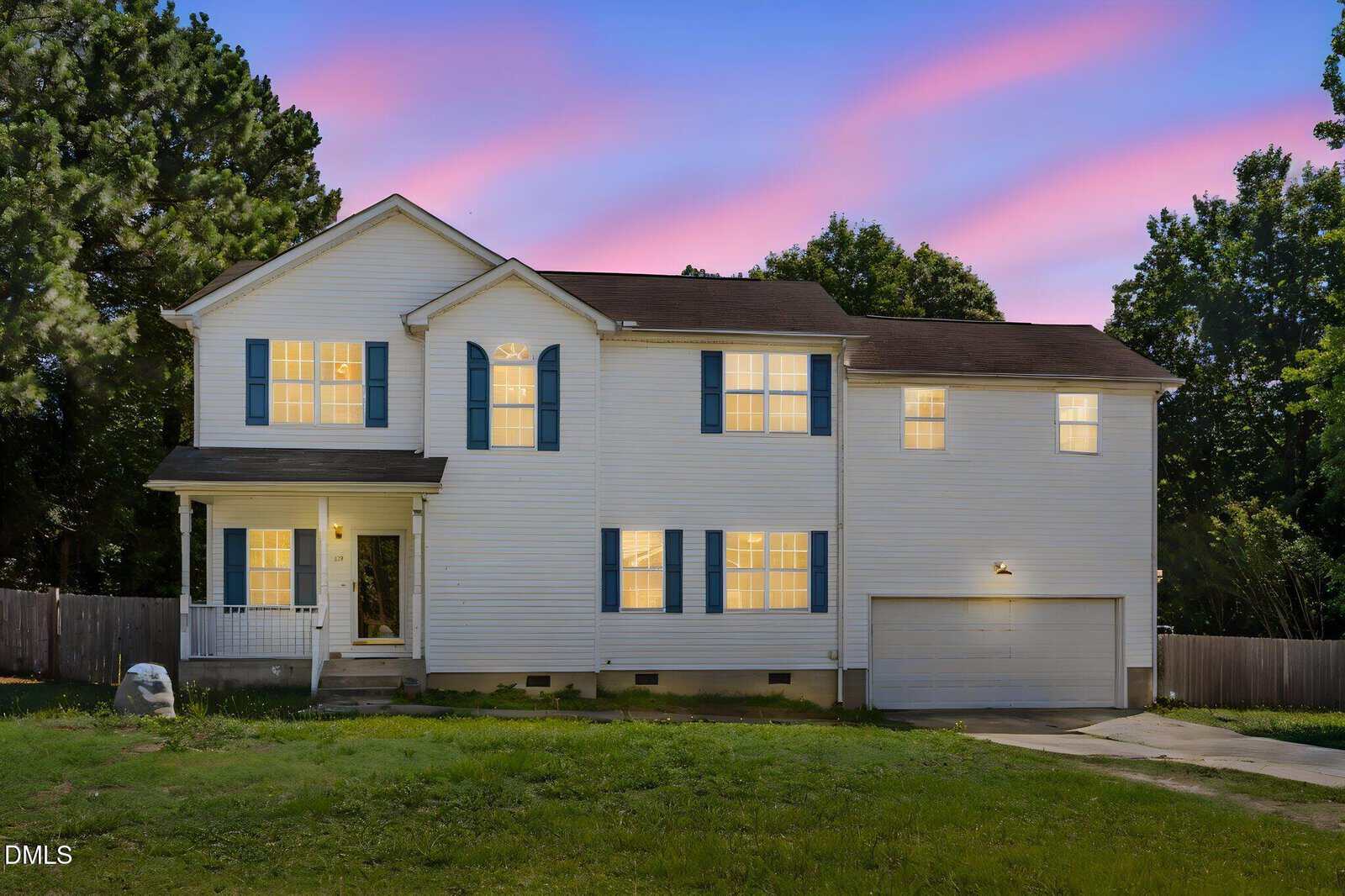Two-story white house with blue shutters, a front porch, and attached two-car garage, illuminated at dusk with a pink and blue sky in the background. Grass lawn and trees surround the property.