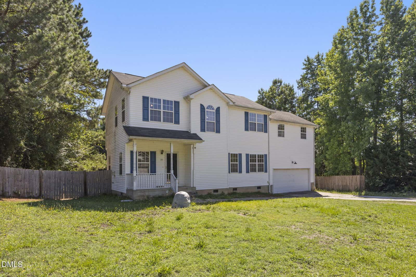 Two-story white house with black shutters, an attached two-car garage, front porch, and large grassy yard. Surrounded by trees and a wooden fence under a clear blue sky.