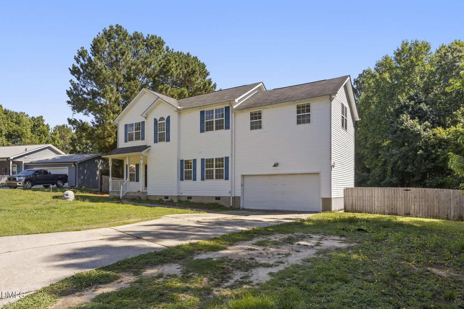 Two-story white house with blue shutters, attached two-car garage, and a grassy front yard. Concrete driveway leads to the garage. Trees and a wooden fence are in the background under a clear blue sky.