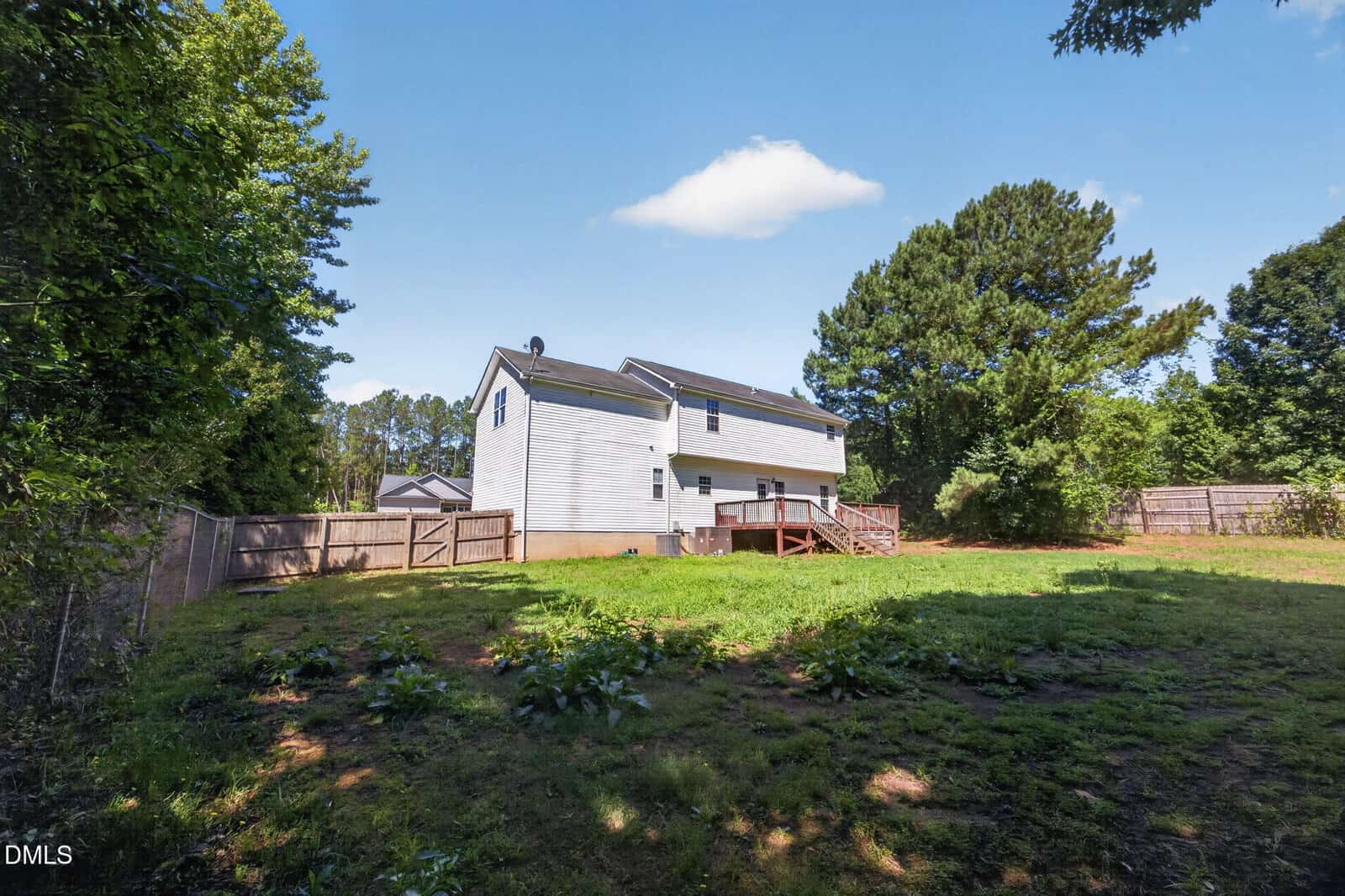 A white two-story house with a wooden deck sits in a large, fenced backyard surrounded by trees under a clear blue sky.