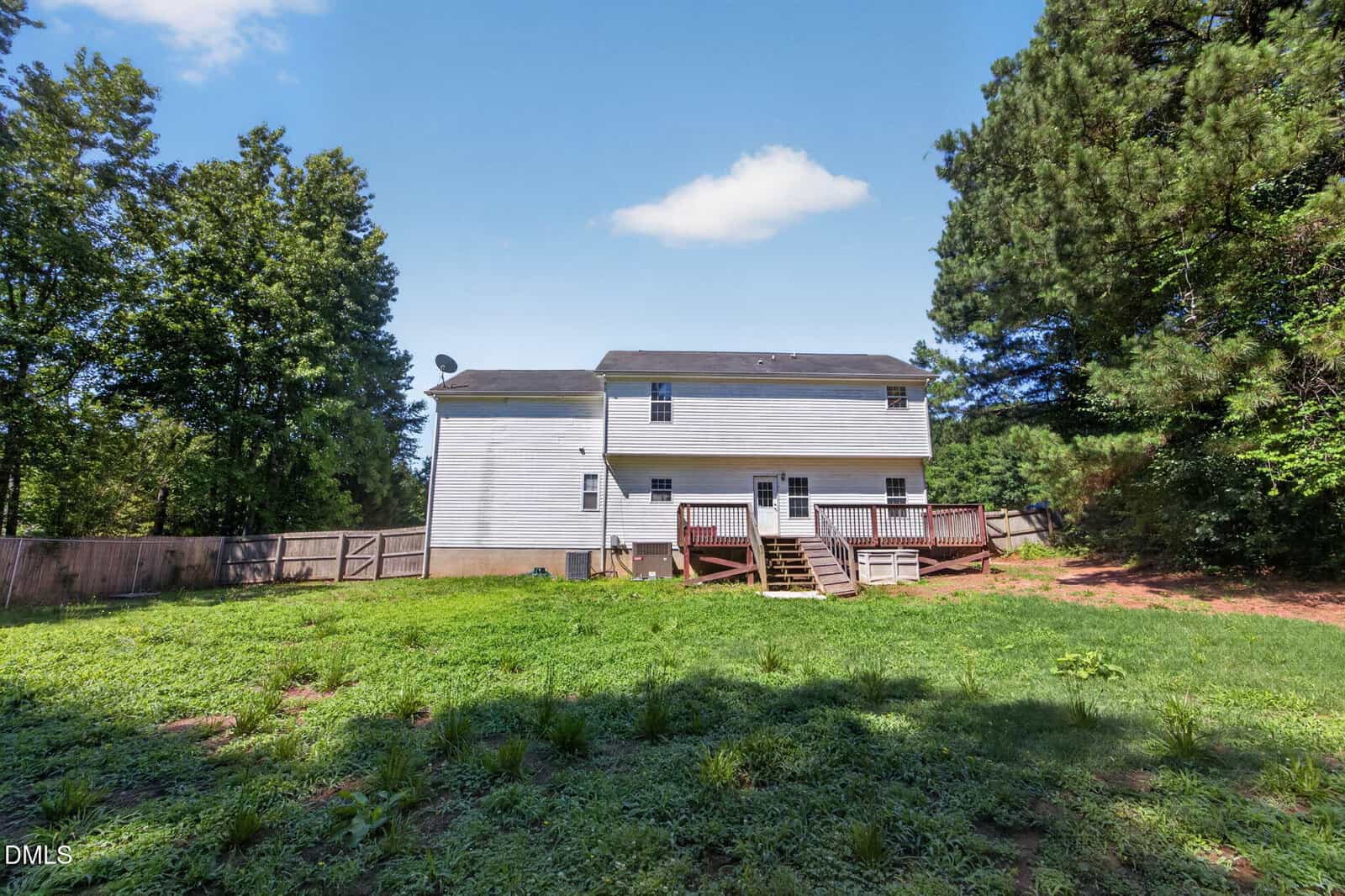 A two-story white house with a wooden deck, stairs leading to a grassy backyard, and surrounded by trees under a blue sky with a few clouds.