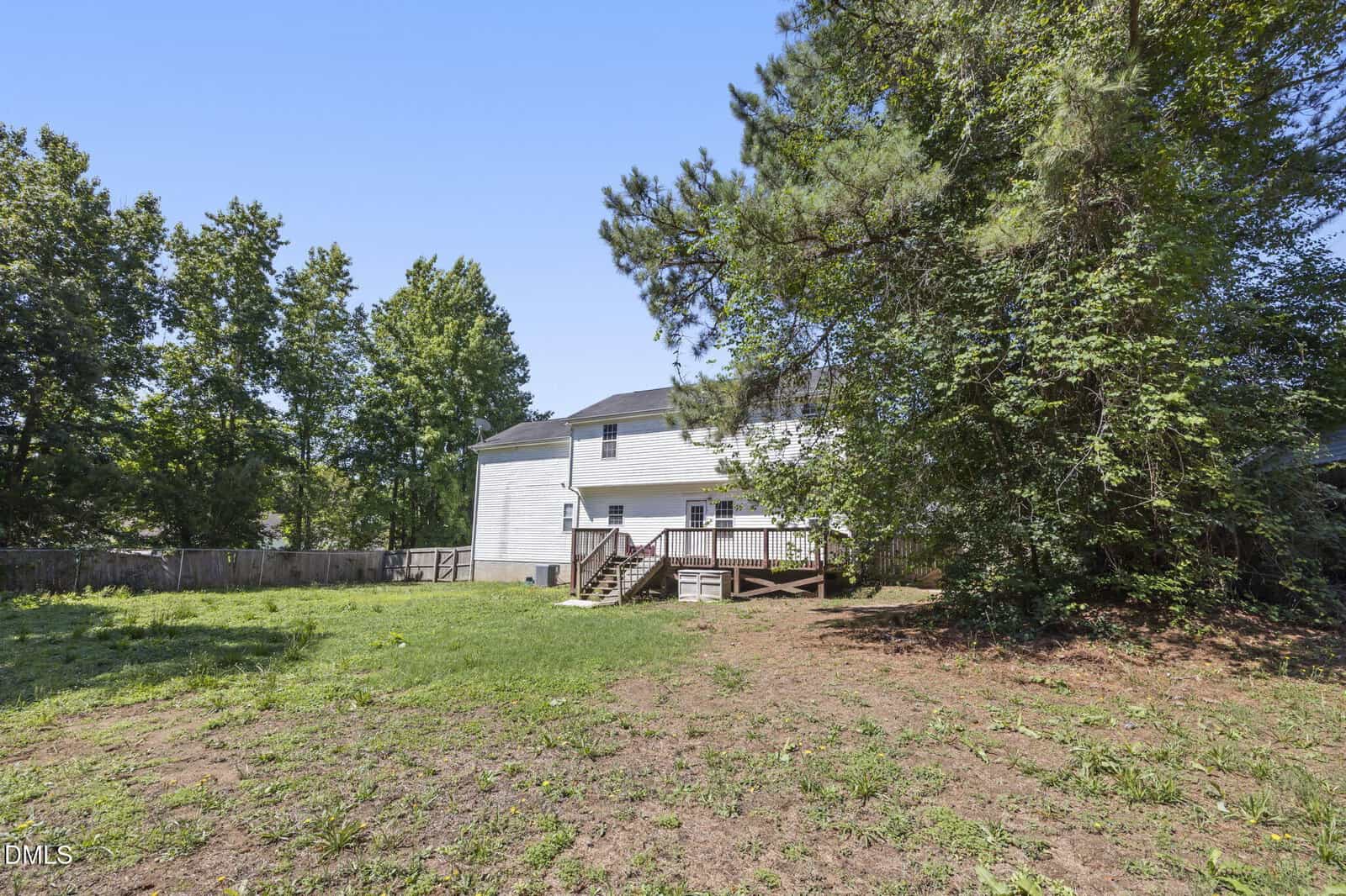 A spacious backyard with patchy grass and dirt, bordered by trees and a wooden fence, features a white two-story house with a raised wooden deck and stairs leading down to the yard under a clear blue sky.