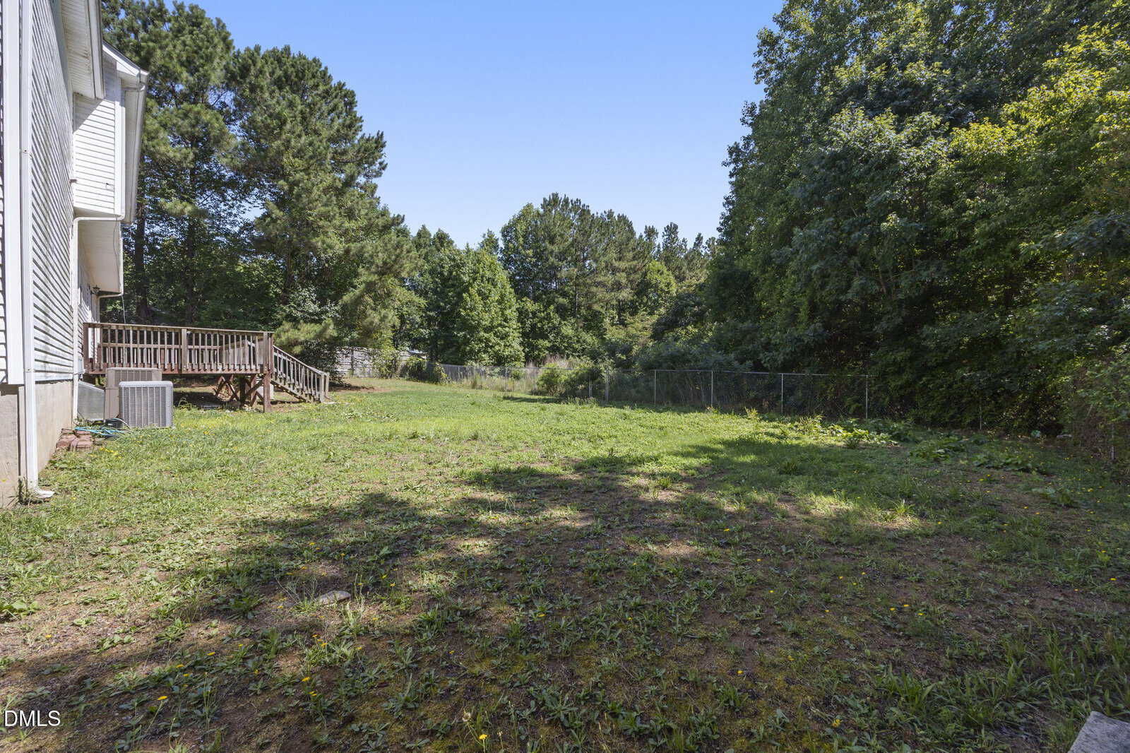 A backyard with sparse grass, a wooden deck attached to a white house on the left, and a fence bordering dense trees and greenery under a clear blue sky.