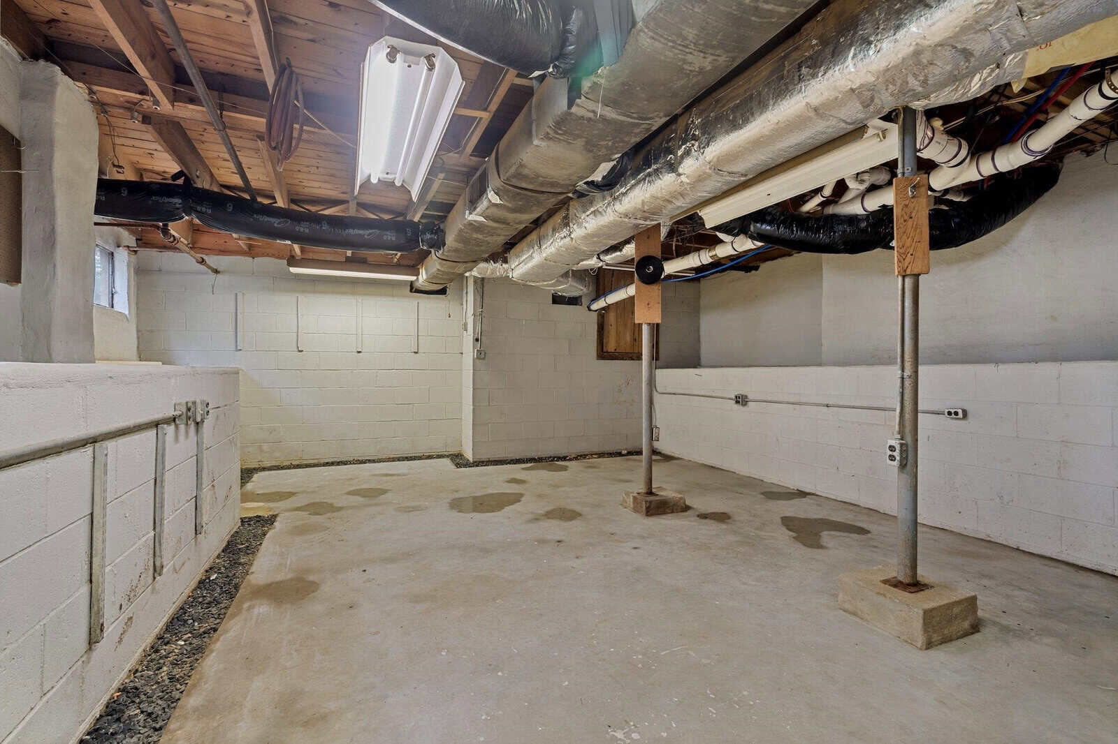 Unfinished basement with exposed pipes, concrete floor, cinder block walls, support poles, fluorescent lights, and some water stains on the floor. Ceiling features visible wooden beams and ductwork.