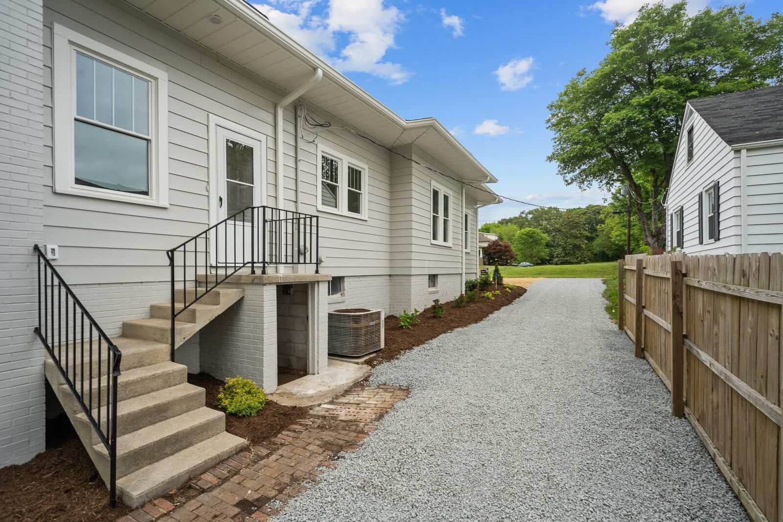 Side view of a white house with a small staircase leading to a door, gravel driveway, wooden fence, central AC unit, and landscaped garden under a partly cloudy sky.