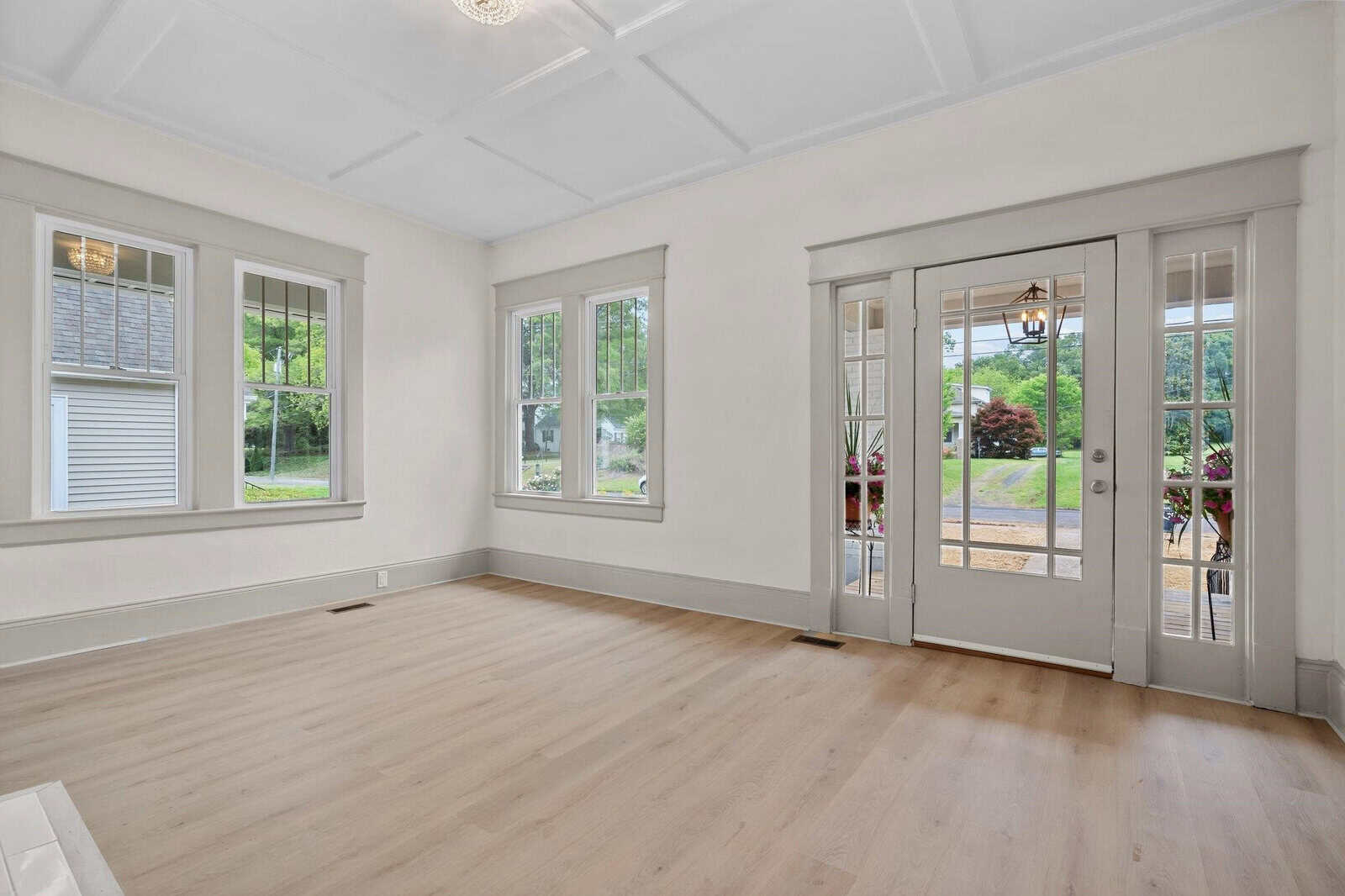 Bright, empty room with light wood flooring, white walls, and large windows. French doors open to a porch, offering views of greenery and a house outside. Natural light fills the space.