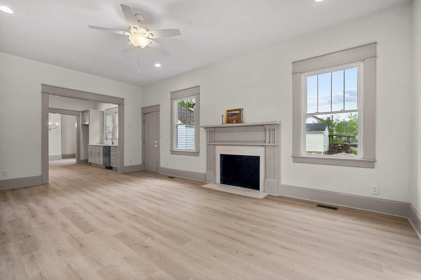 Bright, empty living room with light wood floors, white walls, a ceiling fan, fireplace, and large windows; doorway connects to a kitchen in the background.