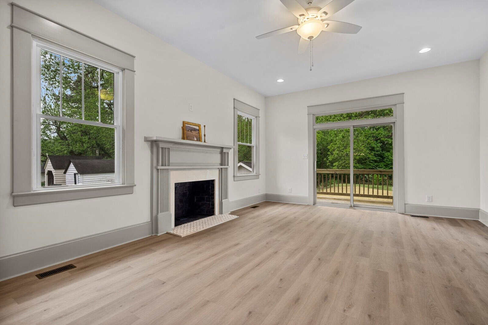 Bright empty living room with light wood floors, a white fireplace, large windows, a ceiling fan, and glass doors leading to a wooden deck with a view of a green yard.