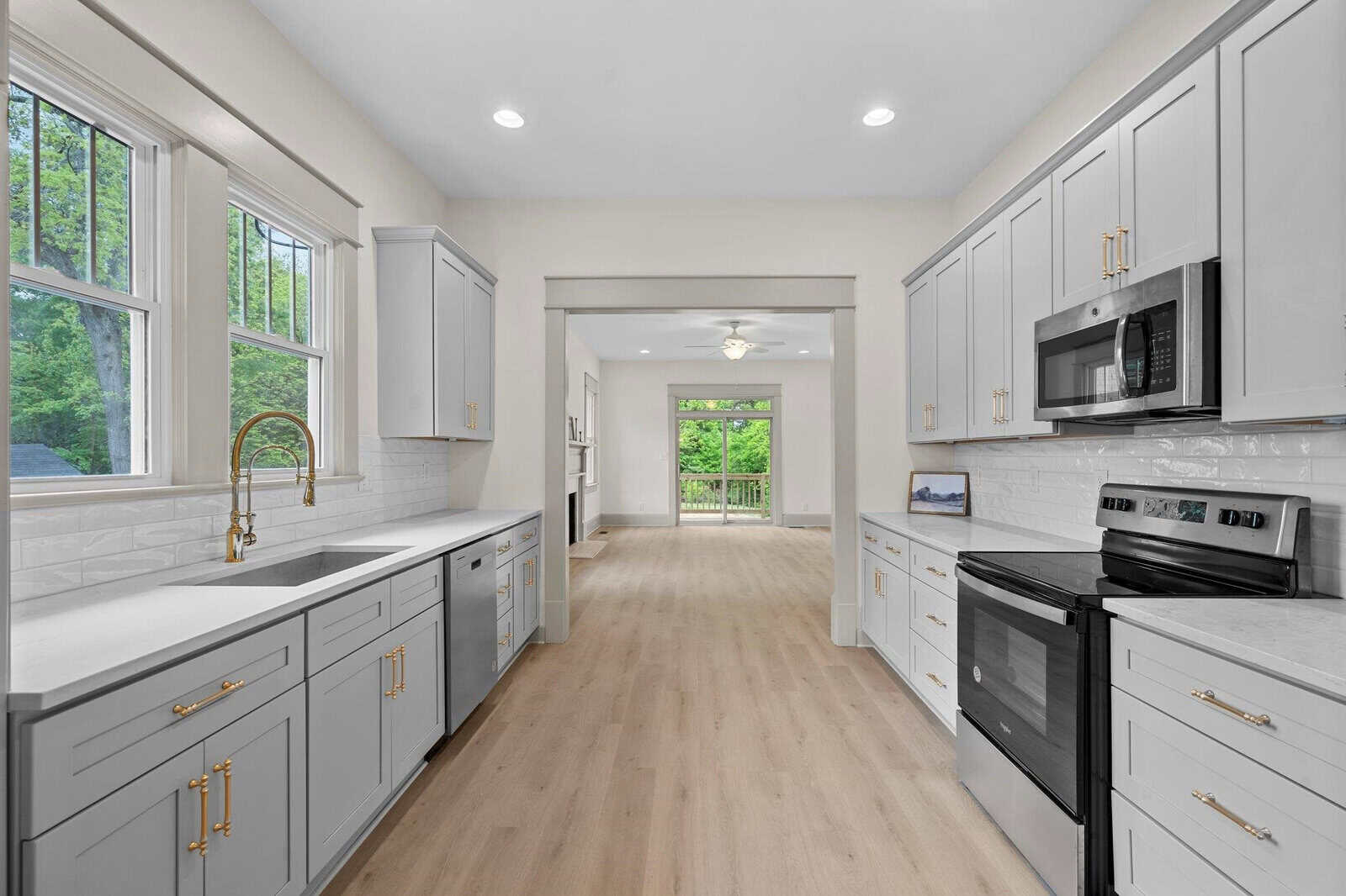 Modern kitchen with gray cabinets, gold hardware, white countertops, stainless steel appliances, farmhouse sink, and light wood floors. Open layout leads to a bright living area with large windows and a ceiling fan.
