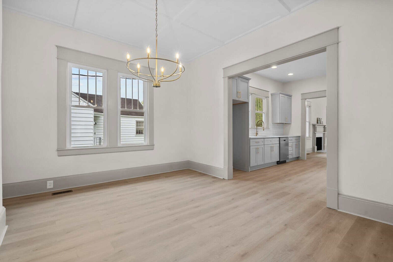 Bright dining room with light wood floors, white walls, a modern gold chandelier, and large windows. Open doorway leads to a white kitchen with cabinets and counters visible.