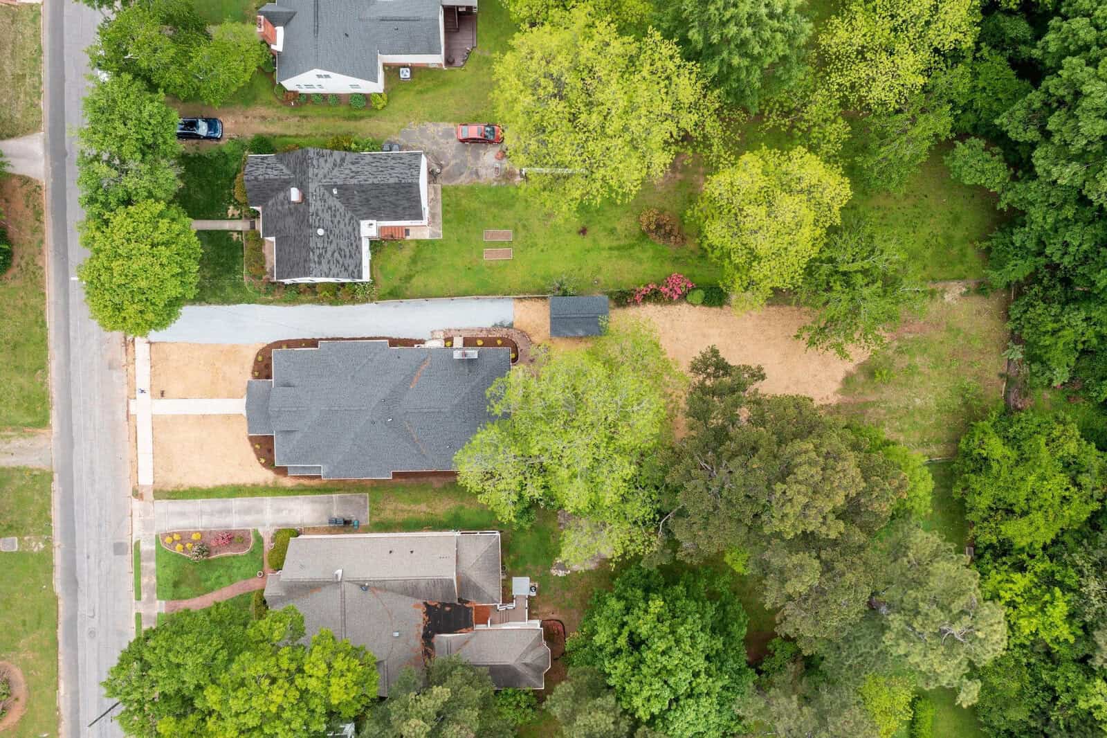 Aerial view of three houses with gray roofs, driveways, green lawns, and surrounding trees. A road runs along the left side, and a large grassy area is visible to the right.