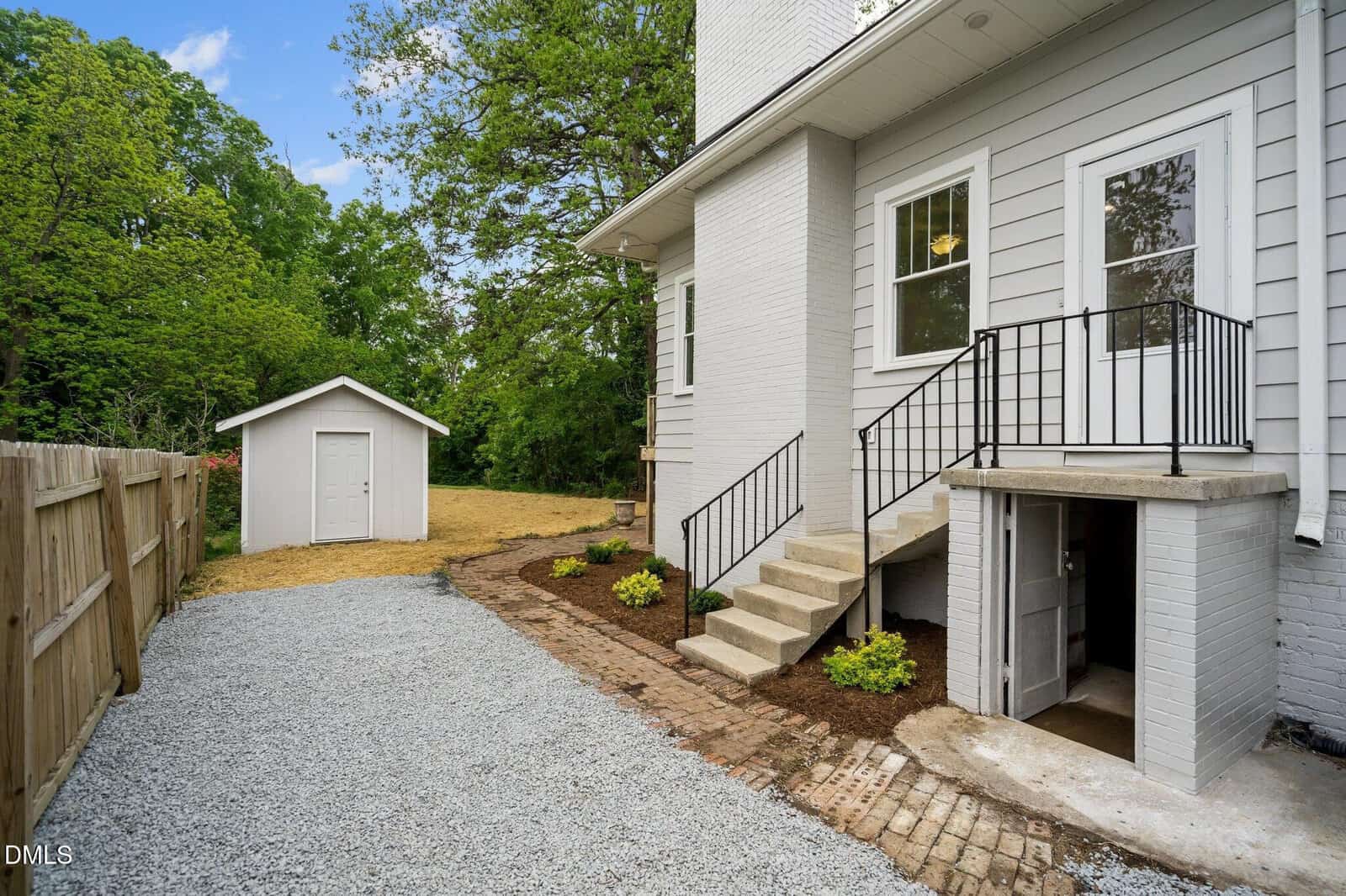 Side view of a white house with a small staircase, a gravel driveway, wooden fence, green trees, and a white shed in the backyard. Small plants are planted along the brick walkway.