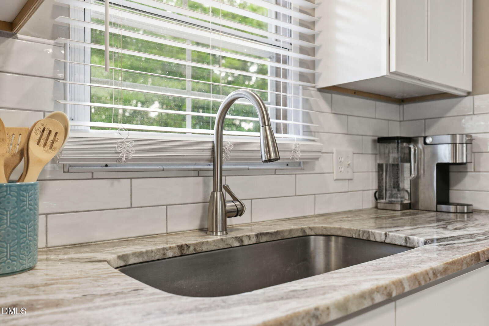 A modern kitchen sink with a stainless steel faucet, marble countertop, utensils in a blue holder, coffee maker, and a window with blinds showing green trees outside.