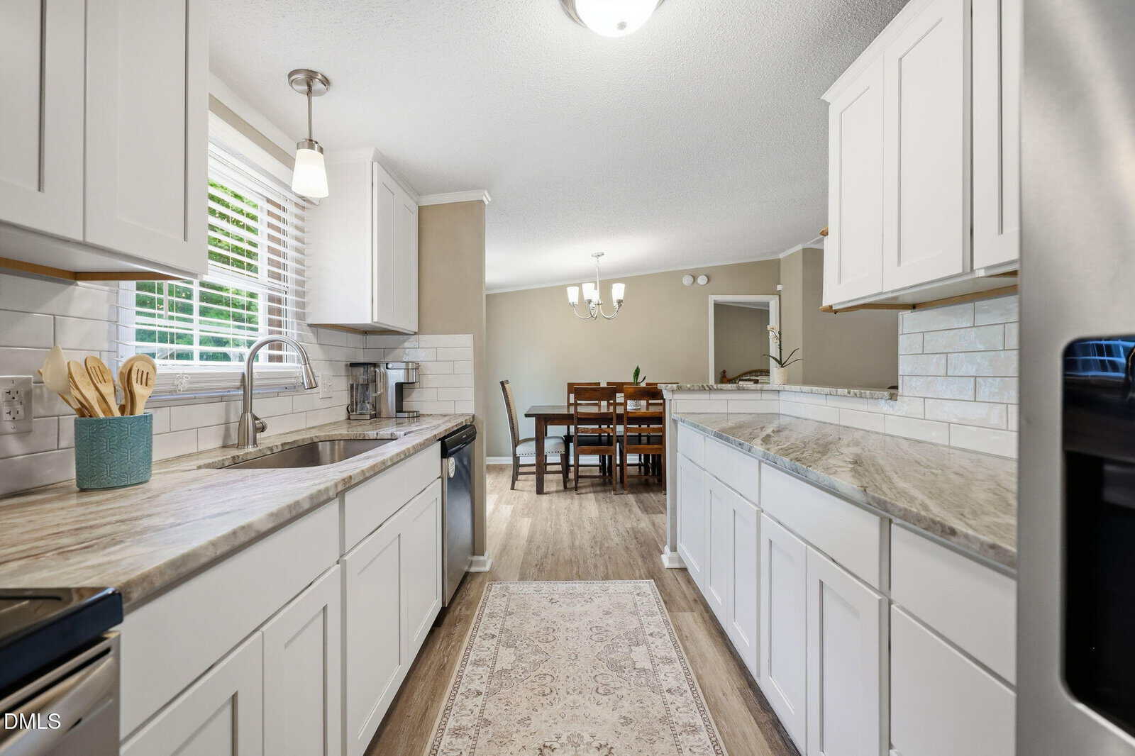 Bright kitchen with white cabinets, marble countertops, a sink under a window, and stainless steel appliances. A dining area with a wooden table and chandelier is visible in the adjacent room.