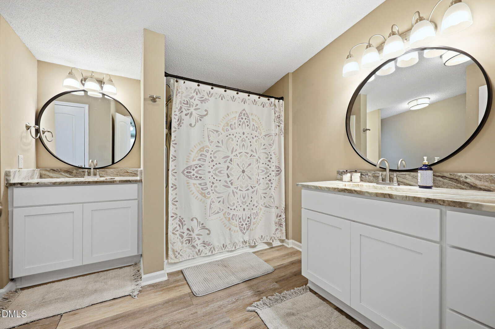 Modern bathroom with two white vanities, marble countertops, and oval mirrors. A shower with a patterned curtain is between the vanities. Beige walls, wood-look flooring, rugs, and overhead lighting create a clean, bright space.