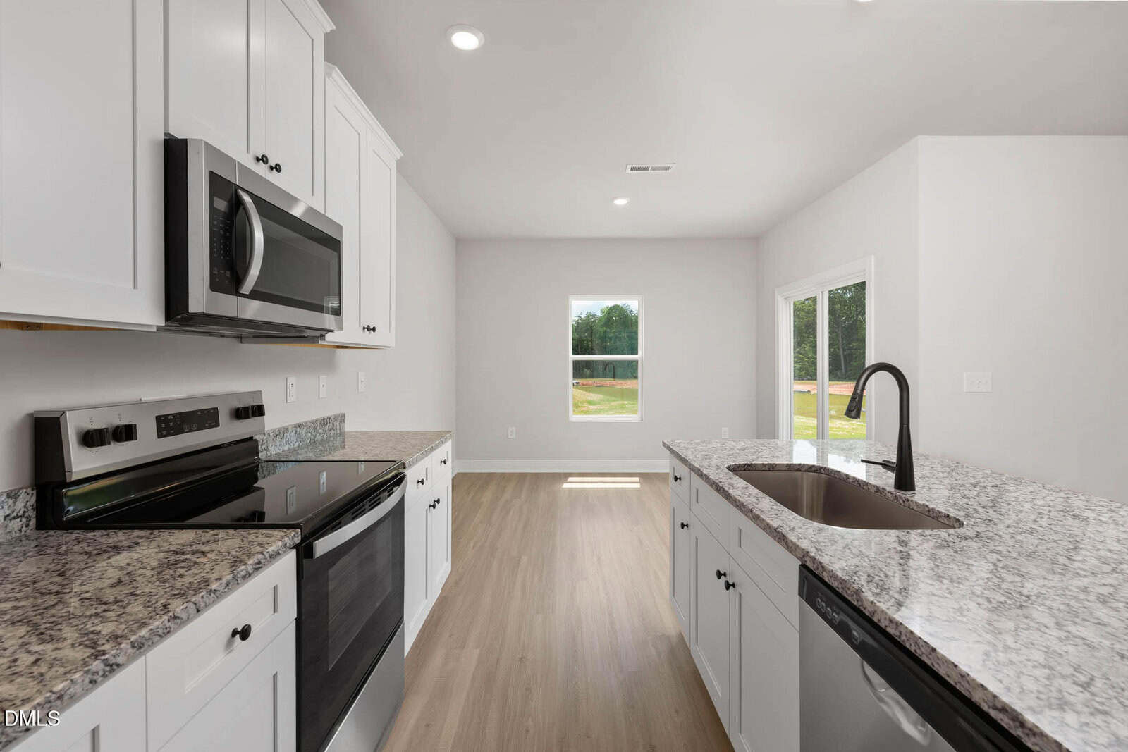 Modern kitchen with white cabinets, granite countertops, stainless steel appliances, and a black faucet. Large window and glass door let in natural light, with a view of greenery outside. Light wood flooring throughout.
