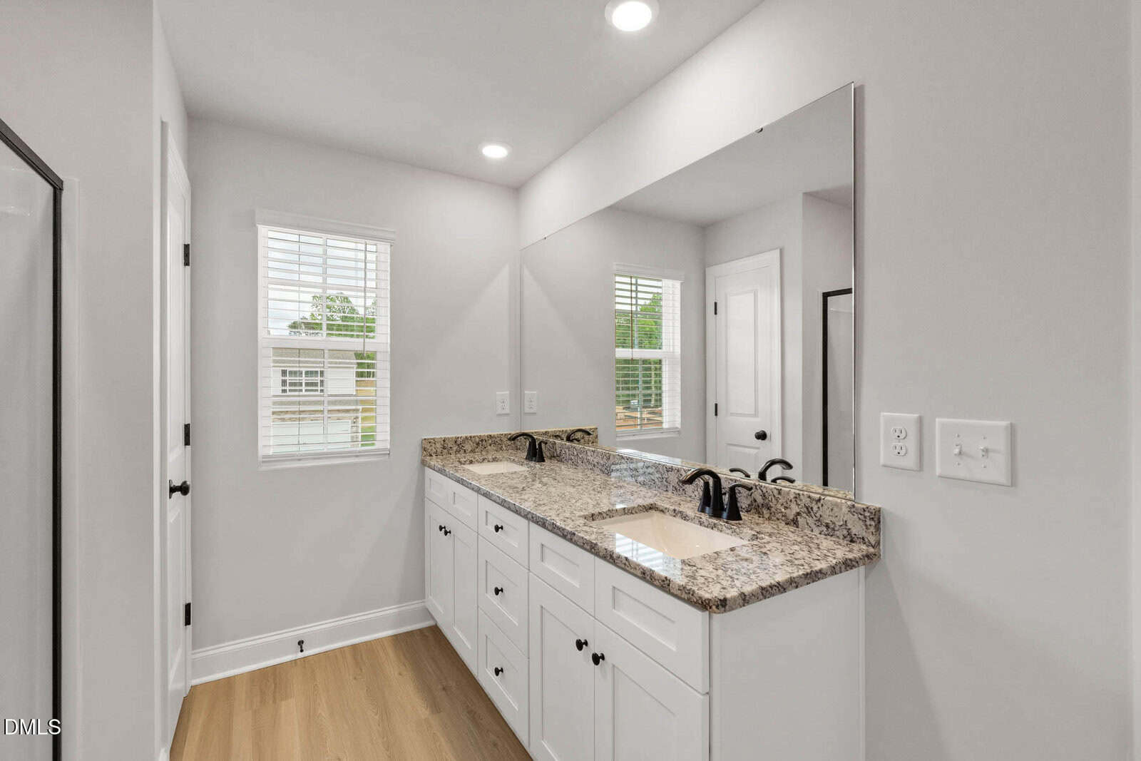 Bright bathroom with double sinks, granite countertop, white cabinets, a large mirror, and two windows. Light wood flooring and neutral walls create an airy, modern feel.