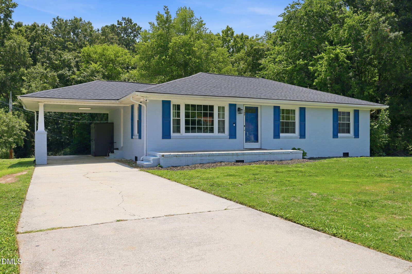 One-story white house with blue shutters, large front window, carport on the left, paved driveway, and green lawn, surrounded by trees under a clear blue sky.