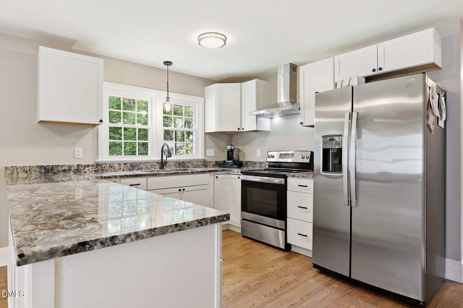 Modern kitchen with white cabinets, stainless steel appliances, granite countertops, a double-door refrigerator, electric stove, sink under a window, and hardwood floors. Bright and clean with ample natural light.