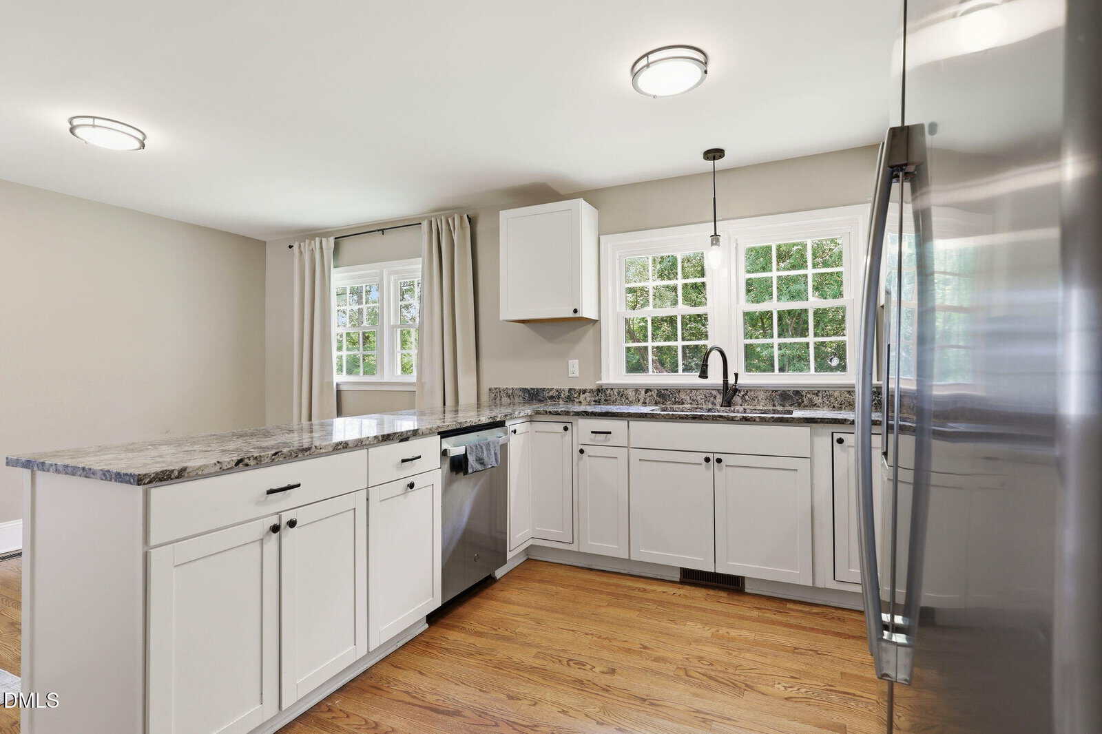 Modern kitchen with white cabinets, stainless steel appliances, granite countertops, wood floor, two large windows with beige curtains, and recessed lighting on the ceiling.