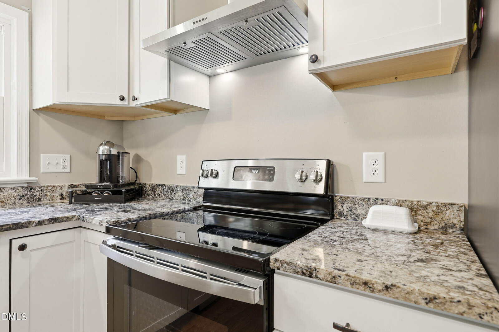 Modern kitchen with stainless steel electric stove, range hood, granite countertops, white cabinets, coffee maker, and a small covered butter dish next to the stove.