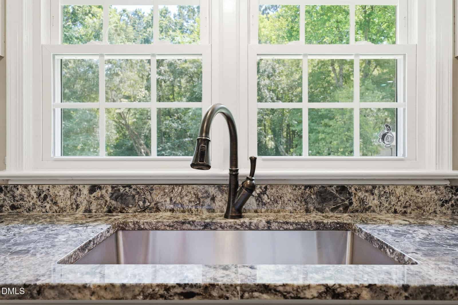 A stainless steel kitchen sink with a dark faucet set in a marble countertop, positioned in front of two large windows showing green trees outside.