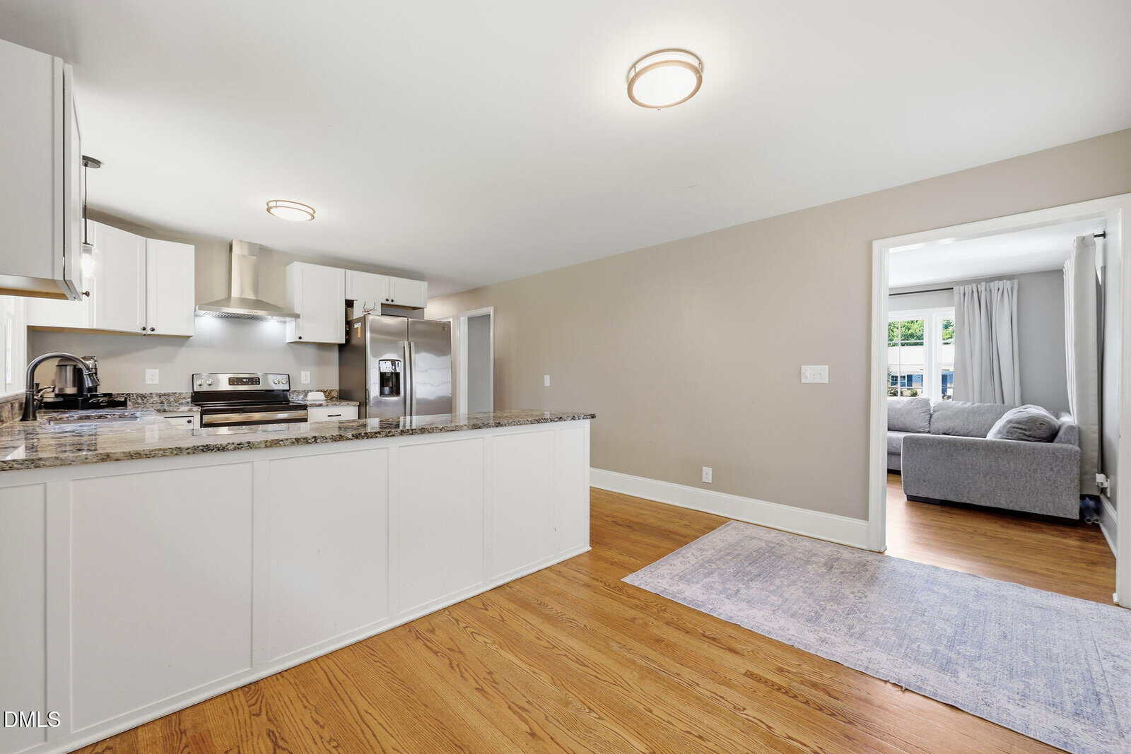 Modern kitchen with white cabinets, stainless steel appliances, granite countertops, and wood flooring. Open doorway leads to a bright living area with a gray sofa and large window. Neutral colors throughout.