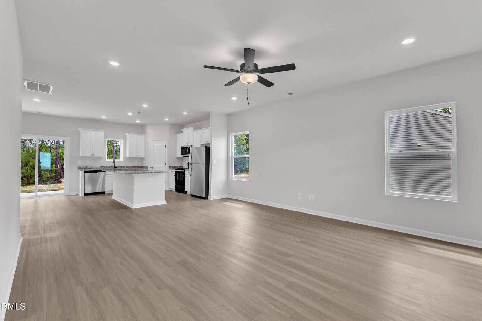 Open-concept living area with light wood flooring, white walls, ceiling fan, recessed lighting, and a kitchen with white cabinets, stainless appliances, and an island. Large windows provide natural light.