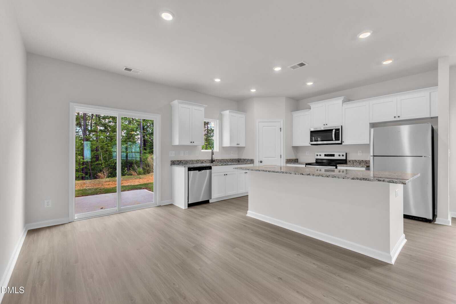 Modern kitchen with white cabinets, stainless steel appliances, and granite countertops. Large sliding glass door opens to a patio with a view of trees. Light wood flooring throughout the open space.