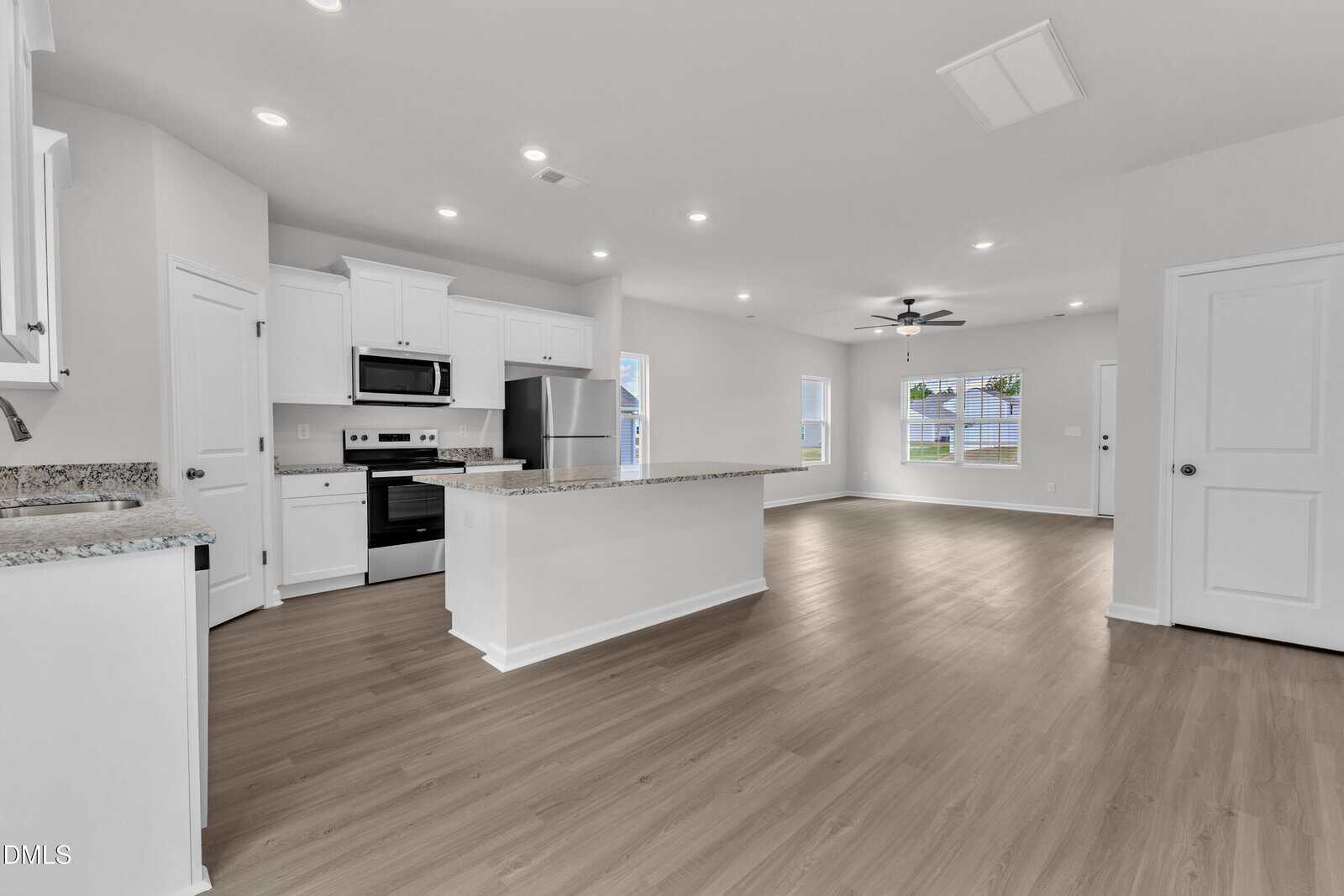 Spacious modern kitchen with white cabinets, stainless steel appliances, and a central island opens to a bright living area with large windows and wood flooring.