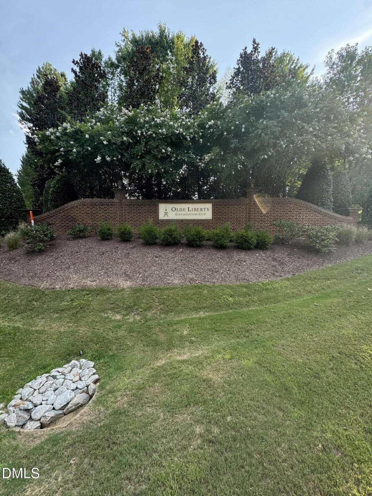 A brick entrance sign for "Olde Liberty Golf & Country Club" sits on a landscaped hill with lush trees and flowering bushes. A small patch of decorative rocks is in the foreground of the green lawn.