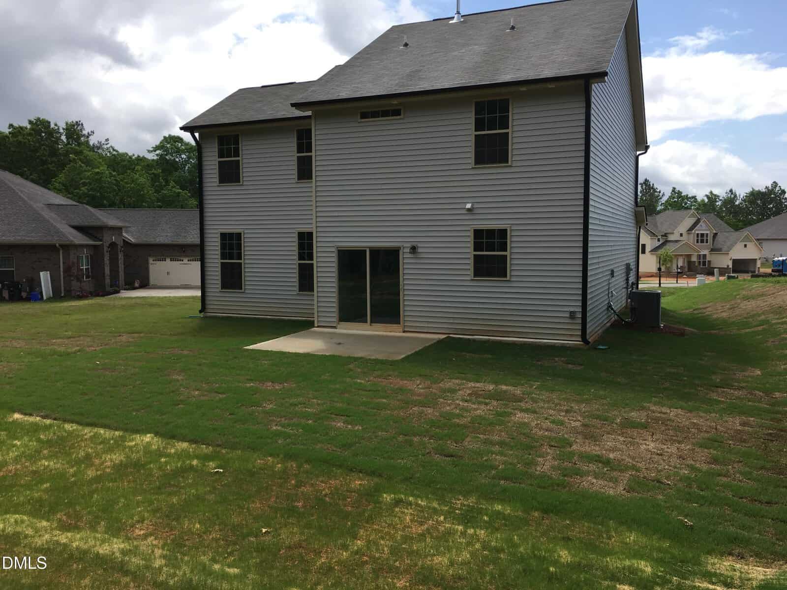 A two-story gray house with a small concrete patio in the backyard, sparse grass patches, and neighboring houses visible on either side under a partly cloudy sky.
