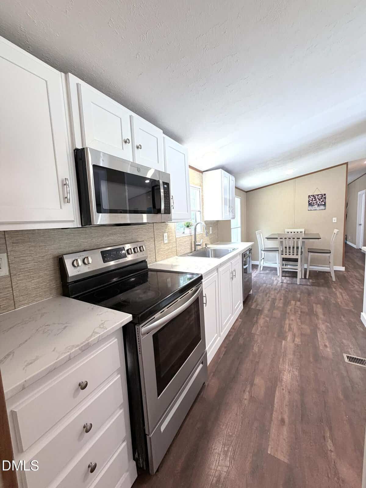 Modern kitchen with white cabinets, stainless steel appliances, and marble countertops. Wood floors lead to a dining area with a table and chairs near large windows, letting in natural light.
