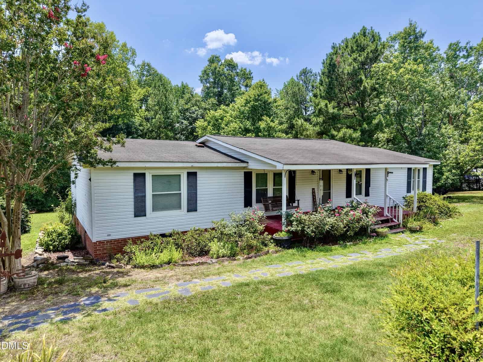 A white, single-story house with a covered front porch, surrounded by green trees and bushes, sits on a grassy lawn under a blue sky at 1702 Mineral Springs Drive in Stem.