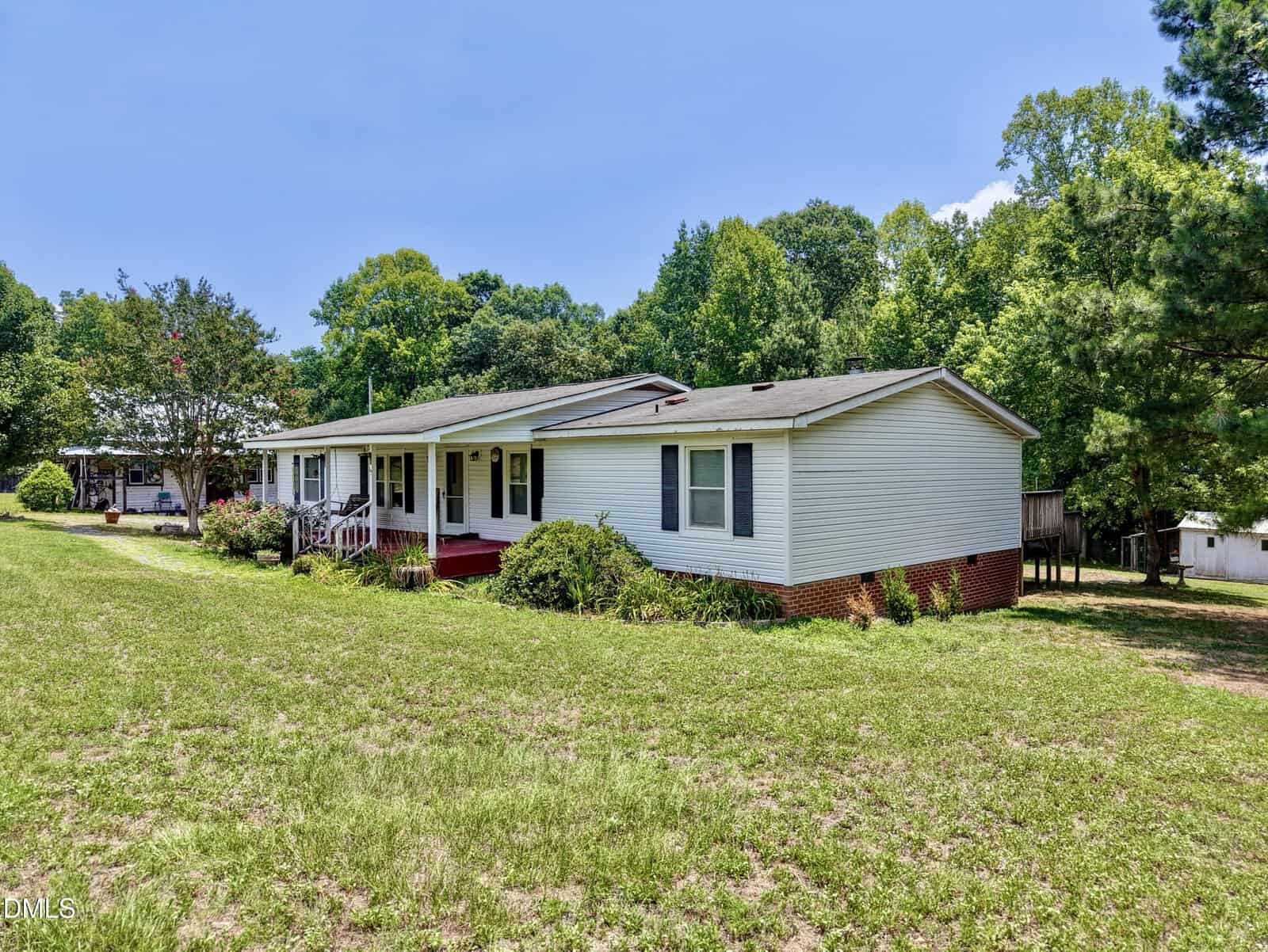 A light blue manufactured home with a red brick foundation, white shutters, and a covered front porch sits on a grassy lot with trees and clear blue sky in the background.